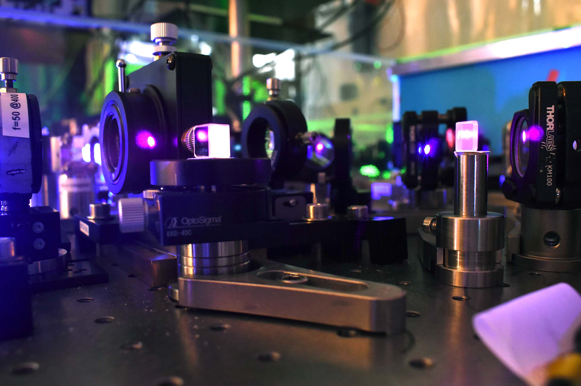 side zoomed-in view of a quantum optics table, with several components locked on steel plate and emitting purple-like laser light.